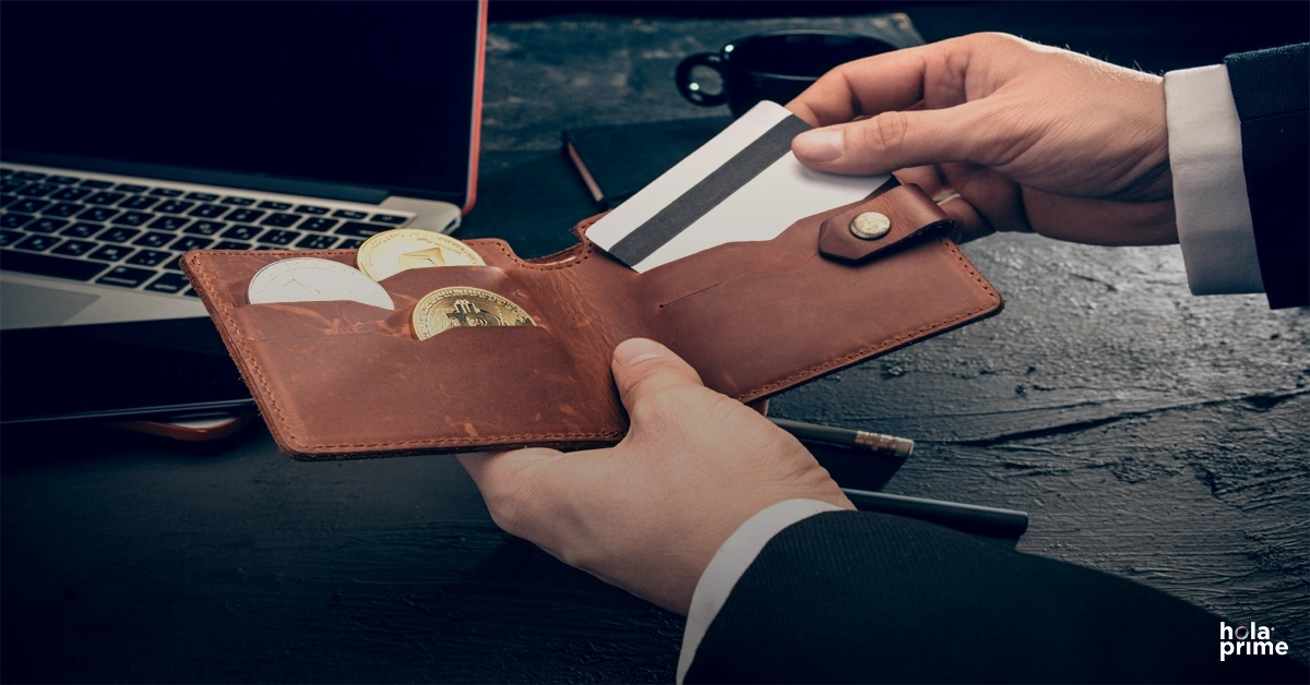 A person in business attire removes a bank card from a brown leather wallet containing cryptocurrency coins while working at a desk with a laptop and coffee cup nearby.